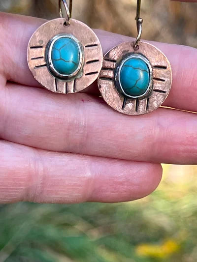 Turquoise stone earrings with bronze or copper setting held between fingers against a blurred natural background.